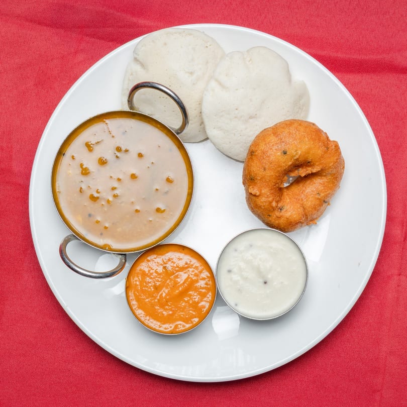 A white plate with idli, vada, sambar, and two chutneys on a red tablecloth.
