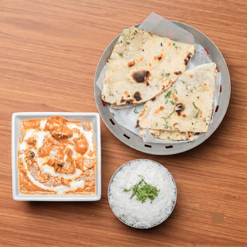 Indian butter chicken, naan bread, and white rice with herbs on a wooden table.
