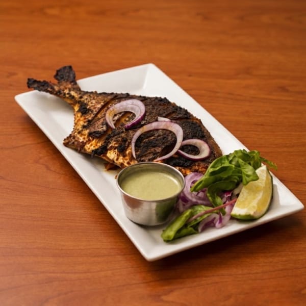 Grilled whole fish with red onion, lime, greens, and a side of green sauce on a white plate.