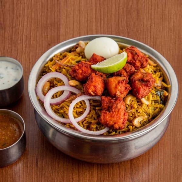 Chicken biryani in a steel bowl, with a boiled egg, lime, and red onions, served with two sauces.