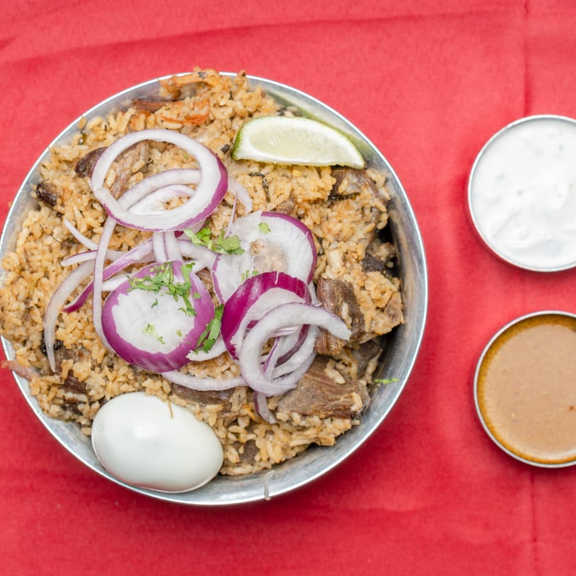 A metal bowl of biryani with red onions, lime, and a boiled egg, served with two sauces.