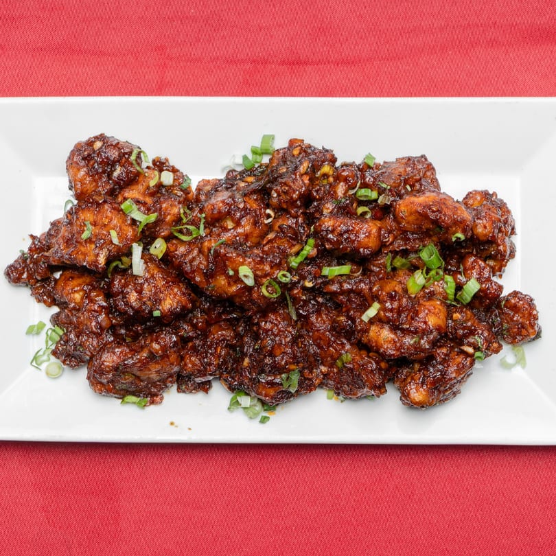 A white rectangular plate filled with dark brown, glazed chicken pieces garnished with chopped green onions.