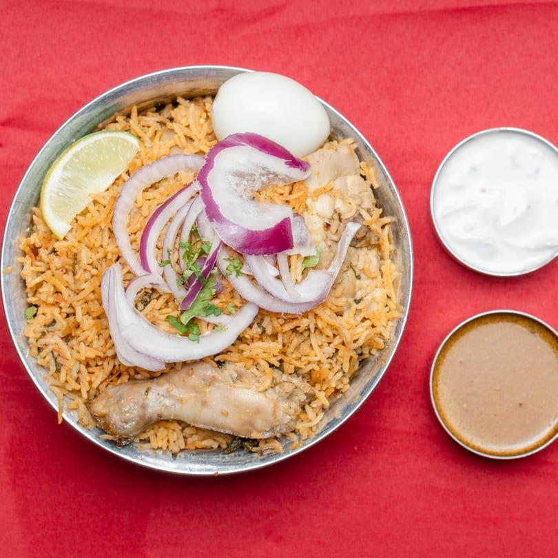 Chicken biryani with sliced red onions, lime, and a boiled egg, served with two sauces.
