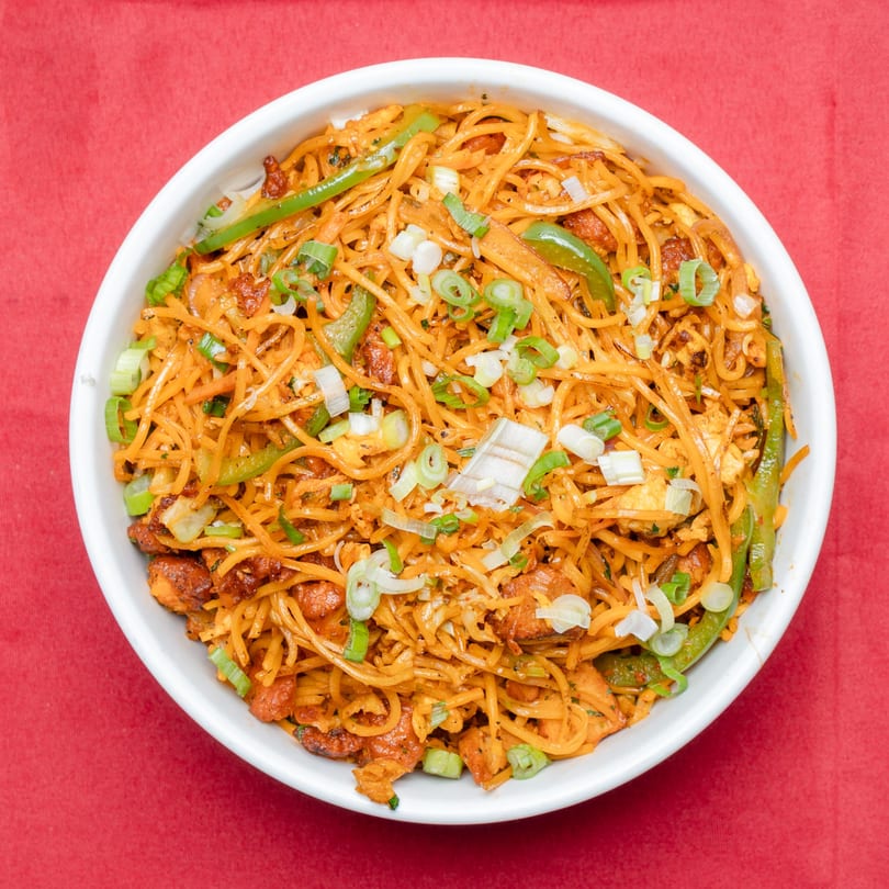 A white bowl filled with chicken noodles, green peppers, and scallions on a red tablecloth.